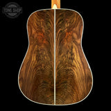 Close-up of the back of an acoustic guitar showing a richly figured burl wood backplate with swirling grain patterns in warm brown and amber tones, set against a solid black background.