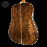 The back of an acoustic guitar featuring dramatic, highly figured burl wood on the soundboard, set against a solid black background with a Tone Shop watermark in the top-left corner.