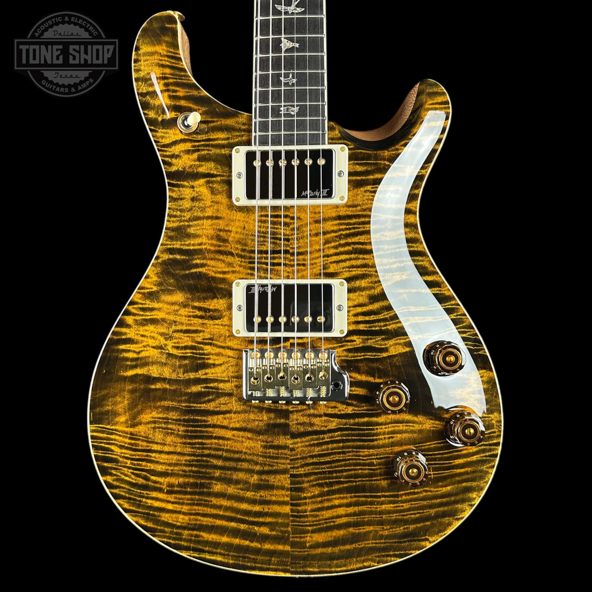 A close-up of an electric guitar body with a glossy flamed maple wood finish in a golden yellow and brown tiger stripe pattern, featuring a white pickguard and gold hardware, set against a solid black background.