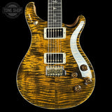 A close-up of an electric guitar body with a glossy flamed maple wood finish in a golden yellow and brown tiger stripe pattern, featuring a white pickguard and gold hardware, set against a solid black background.