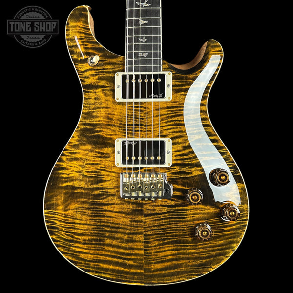 A close-up of an electric guitar body with a glossy flamed maple wood finish in a golden yellow and brown tiger stripe pattern, featuring a white pickguard and gold hardware, set against a solid black background.