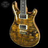 A close-up of an electric guitar body with a dramatic natural figured maple wood finish showing strong golden-brown grain, set against a solid black background. A Tone Shop watermark is visible in the top-left corner.