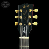 Close-up of a Gibson Les Paul Standard electric guitar headstock, featuring gold tuning pegs, a black finish, and a white Sitari pickup cover, set against a solid black background.