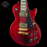 A red electric guitar with a glossy flamed maple top, gold hardware, and a black pickguard, shown against a solid black background with a Tone Shop watermark in the top left corner.