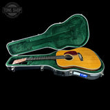 An acoustic guitar with a natural wood finish and a red pickguard, displayed inside an open green-lined hard case on a black background. A Tone Shop watermark is visible in the top-left corner.