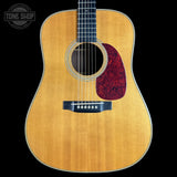 A front-facing acoustic guitar with a natural flamed maple top, dark fretboard, and a red marbled pickguard, shown against a solid black background with a Tone Shop watermark in the upper left.