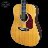 An acoustic guitar with a natural flamed wood top, dark brown sides, and a red pickguard, shown against a solid black background with a Tone Shop watermark in the top left corner.
