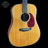 A close-up studio shot of an acoustic guitar body against a black background, showing a natural wood grain top, dark fretboard, and a red marbled pickguard. A Tone Shop watermark is visible in the upper left corner.
