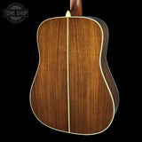 The back of an acoustic guitar with a rich brown wood grain finish and a cream-colored center stripe, shown against a solid black background with a Tone Shop watermark in the top left corner.