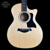 A Taylor acoustic-electric guitar with a natural flamed maple top, black pickguard, and cutaway body, shown against a solid black background. A Tone Shop watermark is visible in the top-left corner.
