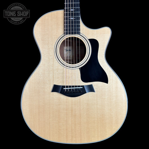 A Taylor acoustic-electric guitar with a natural flamed maple top, black pickguard, and cutaway body, shown against a solid black background. A Tone Shop watermark is visible in the top-left corner.