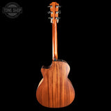 The back view of a wooden acoustic guitar against a solid black background, featuring a warm brown wood grain finish and a Tone Shop watermark in the top left corner.