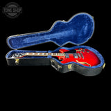 A red electric guitar inside an open black hard case with blue velvet lining, photographed against a solid black background. A Tone Shop watermark is visible in the upper left corner.