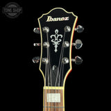 Close-up of the headstock of an Ibanez electric guitar against a black background, showing the black headstock, silver tuning pegs, white Ibanez logo, and white fleur-de-lis emblem.