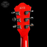 Close-up of the headstock of a red electric guitar against a black background, showing six silver tuning pegs and a white printed sticker with CE markings. A Tone Shop watermark is visible in the top left corner.