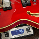 Close-up of a red electric guitar body resting on a digital scale displaying 7.46 lbs, with the scale showing brand Weight Plus.