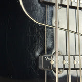Close-up of a black Fender bass guitar body with a black pickup cover, silver metal strings, and visible wear scratches on the glossy finish.
