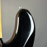 Close-up of the glossy black body of an electric guitar, showing the edge of the neck and a silver tuning peg against a neutral gray background.