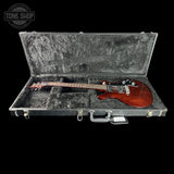 An electric guitar with a reddish-brown body and black headstock, displayed inside an open black hard case with gray foam lining, set against a solid black background.