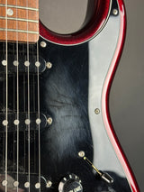 Close-up of an electric guitar body showing a glossy black pickguard with a red edge, silver hardware, and part of the wooden neck on a dark gray background.