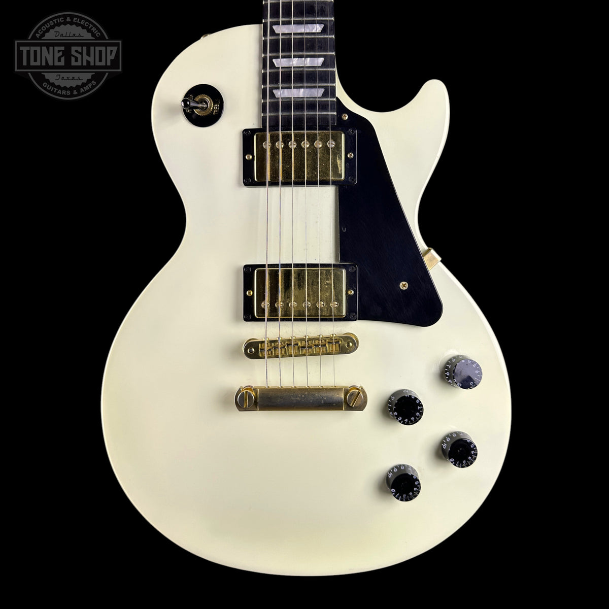 An electric guitar with a glossy off-white body, black pickguard, gold hardware, and black control knobs, shown against a solid black background with a Tone Shop watermark in the top-left corner.