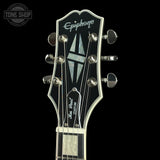 Close-up of the headstock of an Epiphone Les Paul Custom electric guitar against a solid black background. The headstock features a glossy black finish, chrome tuning pegs, a white Epiphone logo, and a geometric inlay.