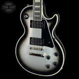 A close-up studio shot of an electric guitar body against a black background, featuring a silver-to-black sunburst finish, white binding, dual black humbucker pickups, chrome hardware, and a visible Tone Shop watermark in the upper left.