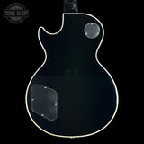 A glossy black electric guitar body, likely a Gibson Les Paul style, shown from the back against a solid black background. It features a white binding edge, a clear circular mounting patch for a pickup cover, and a clear rectangular shield for the control plate.
