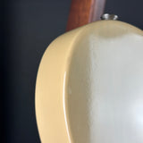 Close-up of a cream-colored guitar body with a glossy finish, showing a portion of the wooden neck and a silver tuning peg against a dark background.
