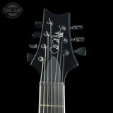 Close-up of a black electric guitar headstock with six tuning pegs and six strings, featuring a white script signature and 'BF' on the backplate, shown against a black background.