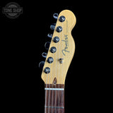 Close-up of the headstock of a Fender Telecaster electric guitar, showing the natural maple wood headstock, six black tuning pegs, and the Fender logo, set against a solid black background.