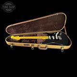 A black Fender Stratocaster electric guitar with a yellow maple neck, displayed inside an open tan hard case with a brown velvet-lined interior, set against a solid black background.