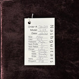 A white paper hangtag with handwritten order details for a Nash guitars body, placed on a close-up of dark brown or very deep espresso wood veneer. The tag details include model S-557, color Blonde, fretboard Maple, neck profile C, neck radius 10, pickups Lollar, Middle Blonde, Bridge Special, and aging Light.