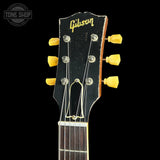Close-up of a Gibson acoustic guitar headstock with a black finish, yellow tuning pegs, and a dark fretboard against a black background.
