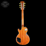 The back view of a classic Gibson Les Paul Custom electric guitar with a warm orange-brown wood finish, shown against a solid black background. The Gibson logo is visible on the headstock and the back body.