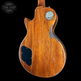 The back of a vintage Gibson electric guitar with a warm brown wood finish, showing a circular Gibson logo badge, a black pickguard, and a worn edge on a black background.