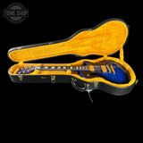 A blue and black electric guitar inside an open black hard case with a bright yellow-orange velvet interior, photographed against a solid black background. A Tone Shop watermark is visible in the top-left corner.