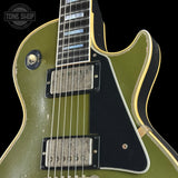 Close-up of a vintage electric guitar body in olive green with a black pickguard, black pickups, gold trim, and blue block inlays on the fretboard. A Tone Shop watermark is visible in the top left corner.