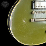 Close-up of the body of an electric guitar with an olive green finish, showing the edge of the body, a gold binding strip, a silver humbucker pickup, and a chrome tailpiece against a black background. A Tone Shop watermark is visible in the top left corner.