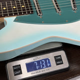 A close-up of a light blue and teal electric guitar resting on a digital scale to show its weight, with the scale displaying 7.13 lbs.