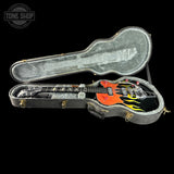 An electric guitar with a black body and orange, yellow, and blue flame graphics, displayed inside an open gray hard case on a black background. A Tone Shop watermark is visible in the top left corner.