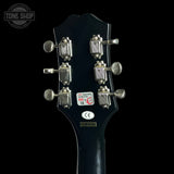 Close-up rear view of a guitar headstock with six silver tuning pegs and stickers, including a Tone Shop logo overlay, set against a solid black background.