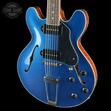 Blue electric guitar with a black pickguard on a black background, featuring the Tone Shop logo.
