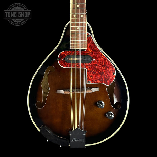 A close-up studio shot of a mandolin against a solid black background. The mandolin features a dark brown sunburst finish, a vibrant red and black marbled pickguard, gold-toned metal hardware, and a black headstock.