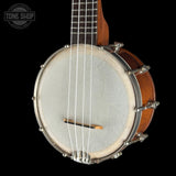 A close-up of a wooden banjo against a solid black background, featuring a warm brown wood body, silver metal hardware, and a light gray drumhead. A circular 'TONE SHOP' watermark is visible in the top-left corner.