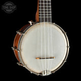 A close-up studio shot of a banjo against a solid black background, showing the wooden body, silver metal rims, white drumhead, neck, and strings. A Tone Shop logo is visible in the top-left corner.