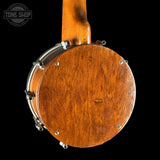 Close-up of the back of a wooden banjo with a warm brown finish, metal lugs, and a neck extending upward against a black background. A Tone Shop watermark is visible in the upper left.
