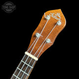 Close-up of the headstock of a Gibson尤克里里 ukulele, showing the brand name 'The Gibson' in white script on a warm brown wood finish, with four tuning pegs and strings against a solid black background.