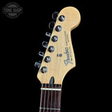 Close-up of a Fender Stratocaster electric guitar headstock in natural maple wood, featuring six black tuning pegs and the blue Fender logo, set against a solid black background.