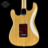 The back of a Fender electric guitar with a natural maple wood finish, a dark tortoiseshell pickguard, and a white curved inlay, shown against a black background.